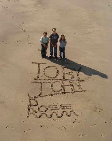 Toby, John and Rose standing by their names written in the sand. On the beach in Whitby, taken from above. It's a glorious sunny day.