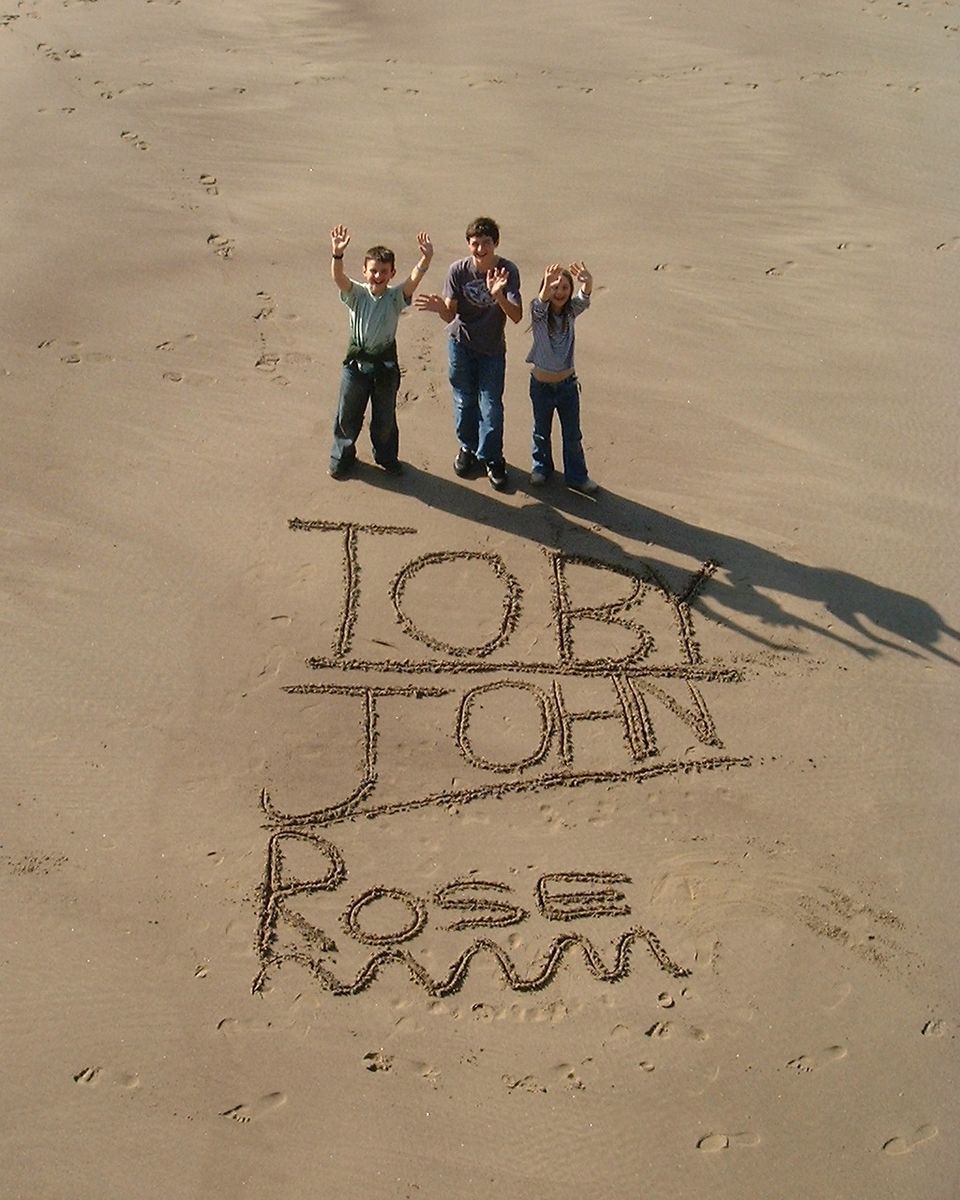 Toby, John and Rose standing by their own names written in the sand. On the beach in Whitby, taken from above. It's a glorious sunny day.